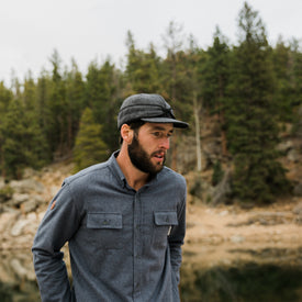 A bearded man stands outdoors near a forested area and rocky lake shore, looking to the side, wearing an Icelantic Stormy Kromer Cap in gray and a button-up shirt.