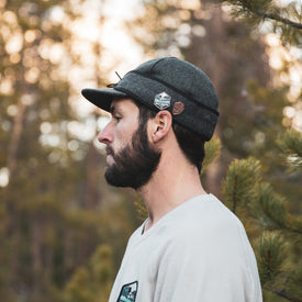 A bearded man in a light shirt stands in profile outdoors, wearing an Icelantic Stormy Kromer Cap, with blurred trees in the background.