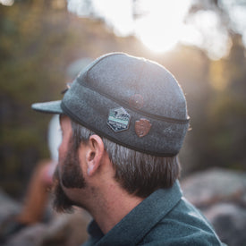 A man with short hair and a beard wears an Icelantic Stormy Kromer Cap in gray, decorated with two outdoor-themed pins. He is outdoors in gentle sunlight, facing away from the camera, with a blurred background of trees and rocks.