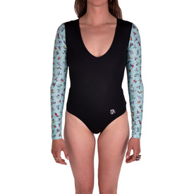 A person is wearing the Icelantic Skier LS One Piece, a black, chlorine-resistant one-piece swimsuit with light blue long sleeves featuring colorful printed designs. The long sleeve swimsuit has a deep scoop neckline, and the face is not visible.