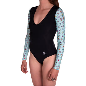 A woman wears the Icelantic Skier LS One Piece, a black long-sleeve swimsuit with light blue patterned sleeves adorned with small colorful birds. The suit features a deep V-neck, built-in UV protection, and a white Icelantic logo on the hip. She stands against a plain white background.