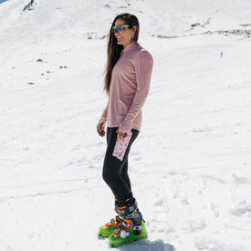 A woman in sunglasses, a pink long-sleeve shirt, and Icelantic Skier Pocket Legging in Black/Pink stands on snow wearing bright green ski boots, with snowy mountains and a blue sky in the background.