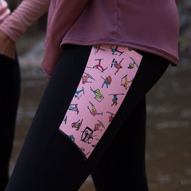 A close-up of a person wearing the Icelantic Skier Pocket Legging in Black/Pink, featuring high-waisted black leggings with a pink side panel adorned with colorful, abstract illustrations of people in various yoga poses. The person pairs the leggings with a mauve top against a blurred background.