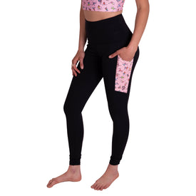 A person is barefoot, standing against a white background and wearing Icelantic's Skier Pocket Legging in Black/Pink—high-waisted leggings made from eco-friendly recycled polyester, featuring a patterned pink side pocket. They’ve paired the leggings with a matching pink crop top.