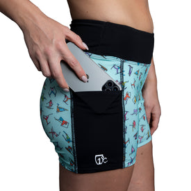 A person places a smartphone into the side pocket of Icelantic's Skier Pocket Short in mint, crafted from recycled fabric and featuring colorful skier illustrations.