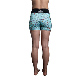 A person stands barefoot facing away, wearing the Icelantic Skier Pocket Short in Mint—a light blue short featuring a playful small shark pattern and a black waistband branded with "ICELANTIC." Made from recycled fabric, these shorts offer a blend of sustainability and fun style against a white background.