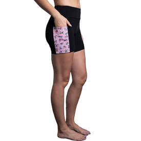 A woman stands sideways barefoot against a white background, wearing the Icelantic Skier Pocket Short in Black/Pink. The shorts feature a pink pocket with a yoga pose print and are crafted from recycled fabric. She has her left hand in the pocket.