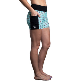A person stands barefoot in profile against a white background, wearing the Icelantic Skier Pocket Short - Mint, a pair of teal and black women's shorts made from recycled fabric featuring a small pocket and printed pattern.