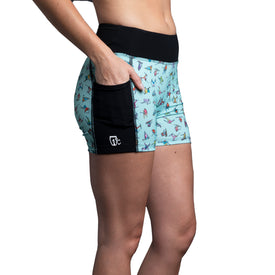 A person wearing Icelantic's Skier Pocket Short in mint—a women's short crafted from recycled fabric with a black waistband, side pocket, small logo, and a pattern of colorful images—stands against a white background.