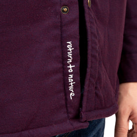 Close-up of a person wearing the Icelantic Sherpa Lined Jacket in dark purple, featuring white embroidered text that reads “return to nature.” Part of the person’s hand and blue jeans are visible, drawing attention to this sustainable piece from Icelantic.