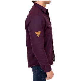 A man stands in profile against a white background, wearing the Icelantic Sherpa Lined Jacket styled as a long-sleeved, dark purple shacket with a patch on the upper arm, paired with blue jeans.