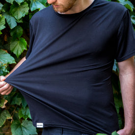 A person outdoors stretches the bottom of their comfortable SoftTECH T - Black from Icelantic Skis with one hand. This shirt, designed with advanced wicking technology, features a small white “CELANT” tag. Green leafy plants are in the background, and the person's face is partially visible.