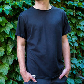 Wearing the Icelantic Skis SoftTECH T in black and dark pants, a person stands with hands in pockets before a leafy green wall.