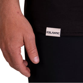 A close-up of a person’s left hand resting by their side, next to a black garment with a white Icelantic Skis label sewn near the hem, highlights the SoftTECH T - Black t-shirt, designed with advanced wicking technology for all-day freshness.