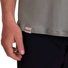 A close-up of a person’s left hand resting near the hem of the Icelantic SoftTECH T - Greenish, a gray, comfortable t-shirt with an antimicrobial CELANTIC tag, worn above black pants.