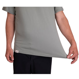 A person wearing the Icelantic SoftTECH T - Greenish stretches the side of the light greenish, comfortable t-shirt outward with their right hand, demonstrating the fabric’s flexibility. Only their torso and arms are visible, paired with dark pants.
