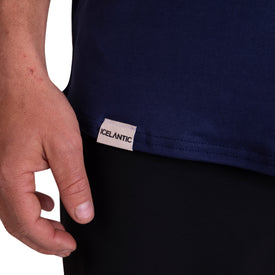 A close-up shows a person's left hand next to the hem of the Icelantic SoftTECH T - Navy, featuring a small white Icelantic tag. Crafted with advanced wicking fabric for unparalleled comfort, this t-shirt is paired with black pants.