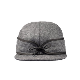 A gray wool Stormy Kromer Cap by Icelantic, featuring a short brim, black trim, and a black cord tied in a bow at the front, offers both warmth and timeless style for winter.