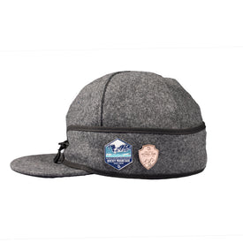 The Icelantic Stormy Kromer Cap is a gray wool hat featuring a side zipper and two embroidered patches: one depicting mountains labeled "Rocky Mountain" and another tan, shield-shaped patch with text. Shown from the side against a white background, this cozy winter cap echoes the timeless Stormy Kromer style.