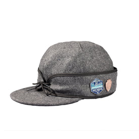 The Icelantic Stormy Kromer Cap is a grey wool hat with a short brim, black trim, and two patches—a blue and white hexagonal patch and a small tan shield patch—shown against a white background.