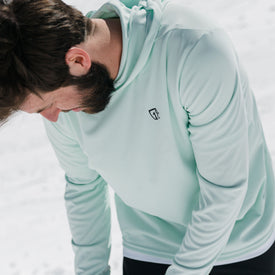 A man with brown hair and a beard, wearing the Icelantic UV Sun Hoodie in Clearly Aqua (UPF 50), looks down while standing outside on a snowy day.