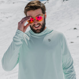 A man wearing the Icelantic UV Sun Hoodie in Clearly Aqua, offering UPF 50 sun protection, adjusts his reflective sunglasses while standing in a snowy outdoor setting.
