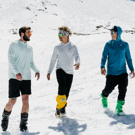 Three men in snow gear and boots walk and smile together on a snowy mountain under a clear, sunny sky, staying comfortable in their Icelantic UV Sun Hoodie - Clearly Aqua, which provides UPF 50 sun protection.