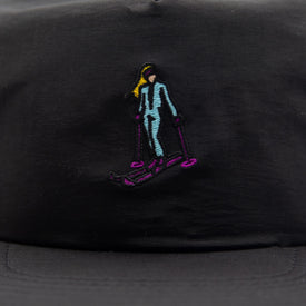 The Hey Girl Nylon Hat by Icelantic Skis is a black cap featuring an embroidered skier in light blue attire, with purple skis and poles, and yellow hair—designed with a floatable bill, making it perfect as a summer hat.