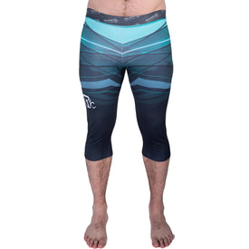 A person is standing barefoot on a white background wearing the Icelantic 22/23 Men's WikMax Baselayer 3/4 Bottom compression leggings in teal and navy blue with a geometric pattern.