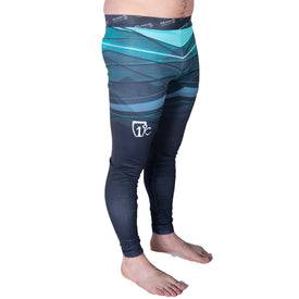 A man stands barefoot on a white background wearing Icelantic's 22/23 Mens WiKmax Baselayer Full Bottoms, which showcase a teal and black geometric pattern and feature a white logo on the left thigh. These baselayer leggings are crafted from innovative WIKMAX fabric.