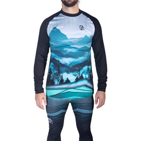A person stands against a white background wearing Icelantic's 22/23 Mens WikMax Baselayer Top from the Pioneer Collection, featuring a striking mountain and forest landscape design in blue and green tones, crafted from innovative WIKMAX fabric.