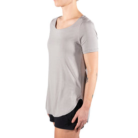 A person wears the Icelantic Skis Eco Bamboo T in Sand—a lightweight, short-sleeve bamboo shirt with a scoop neckline, relaxed fit, and subtle scalloped hem—paired with black shorts while standing sideways against a white background.