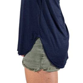 Wearing the Icelantic Skis Eco Bamboo T in navy—a lightweight bamboo shirt with a rounded, side-slit hem—paired with green denim shorts featuring frayed edges. The photo captures the left side of the lower torso and upper thigh.