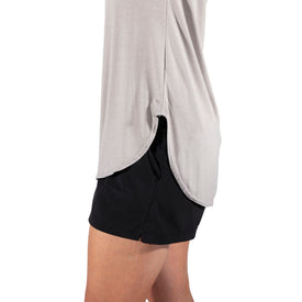 A person wearing the Icelantic Skis Eco Bamboo T in Sand—a light gray, lightweight bamboo shirt with a scalloped hem—paired with black shorts, shown from the shoulders to mid-thigh against a white background. This odor-resistant top combines comfort and style for everyday wear.